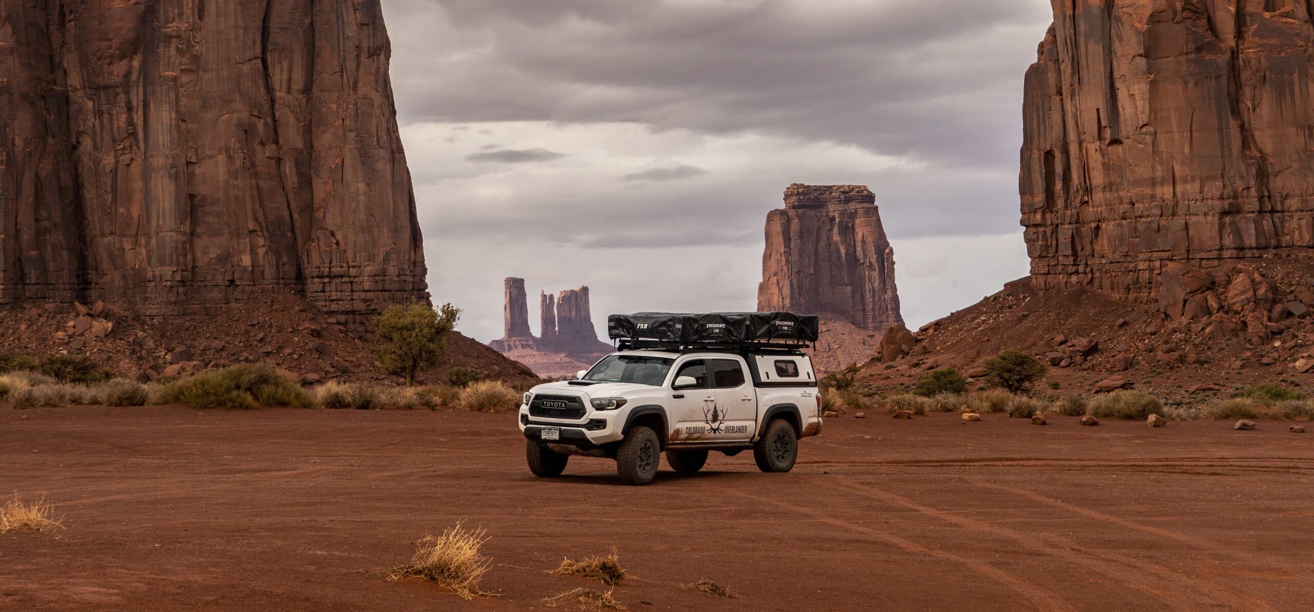 A photo of the ultimate overlanding adventure with our rugged white overland truck set against a dramatic red desert backdrop. Towering rock formations frame the scene, creating a striking contrast against the gray sky adorned with gentle clouds. This purpose-built overland vehicle, complete with roof-top tents, stands ready to traverse the vast and awe-inspiring desert terrains.