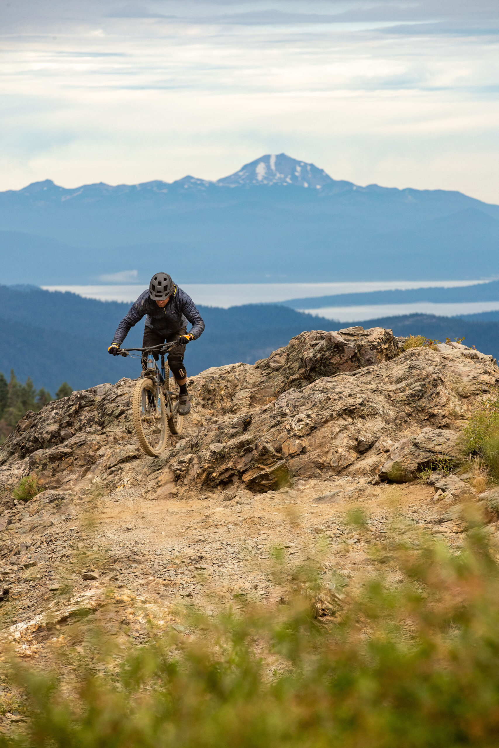 This photo shows a mountain biker going off a rocky jump with mountains in the background.