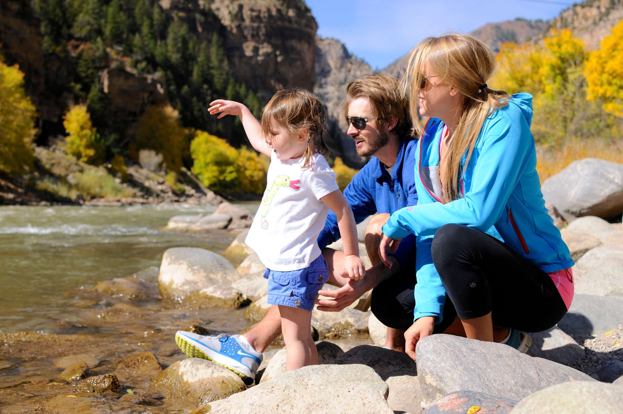In this captivating scene, a family comprising a mother, father, and toddler enjoy a leisurely moment beside the river in Glenwood Canyon. Their gaze is fixed towards the flowing river, as if absorbing the surrounding beauty. Behind them, a backdrop of lush forest and distant canyon walls completes the picturesque setting.