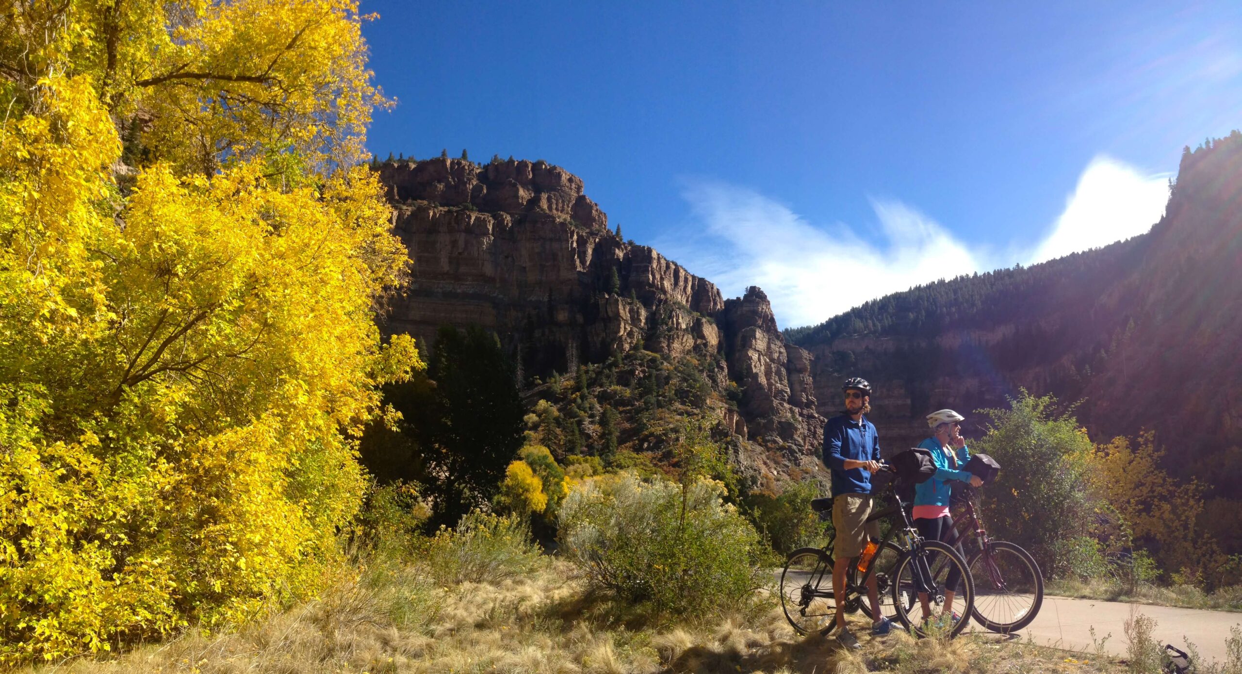 This image features two bikers taking a pause on the Glenwood Canyon Trail. Enveloped by the breathtaking surroundings, they appear to be immersed in the scenic beauty. The trees showcase vibrant fall foliage, harmonizing with the rugged rocky canyon walls.