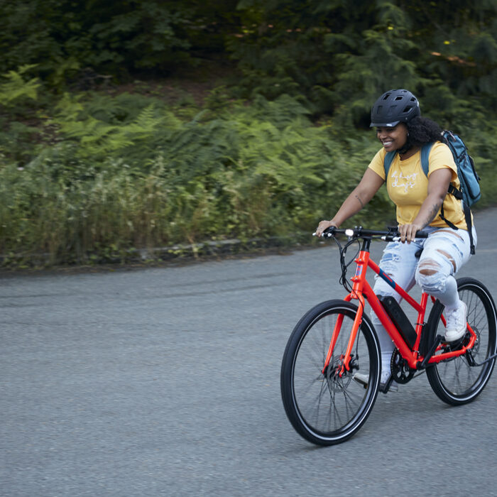 Image of a woman riding a red Rad Mission E-bike- She is wearing jeans and a t-shirt along with her helmet and backpack. The pavement is flat and offers green forest background.